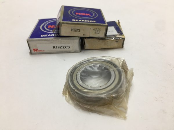 NSK R18ZZC3 deep groove ball bearing in plastic wrap, with stacked NSK bearing boxes behind it